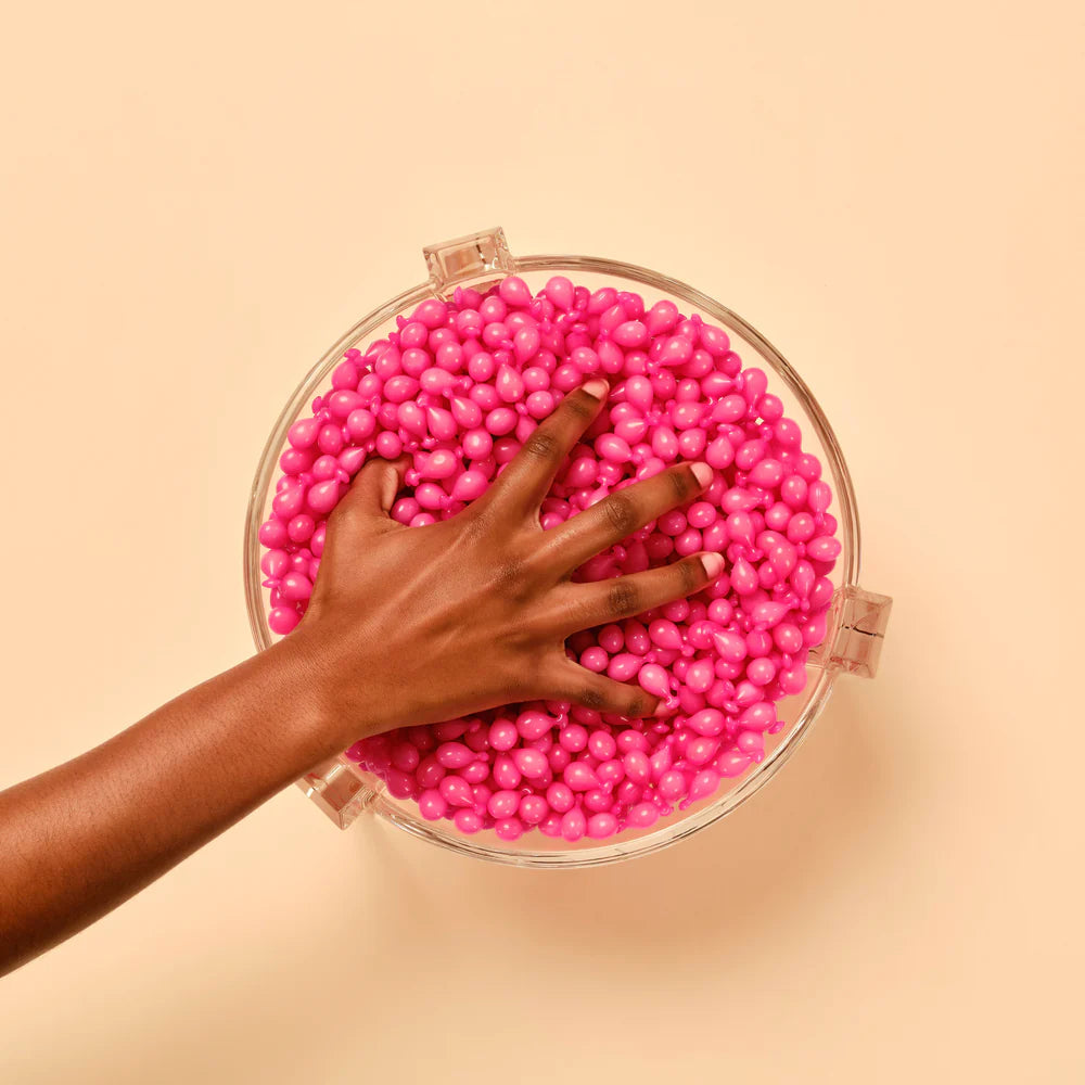 Hand reaching into bowl of bright pink wax beads on beige background