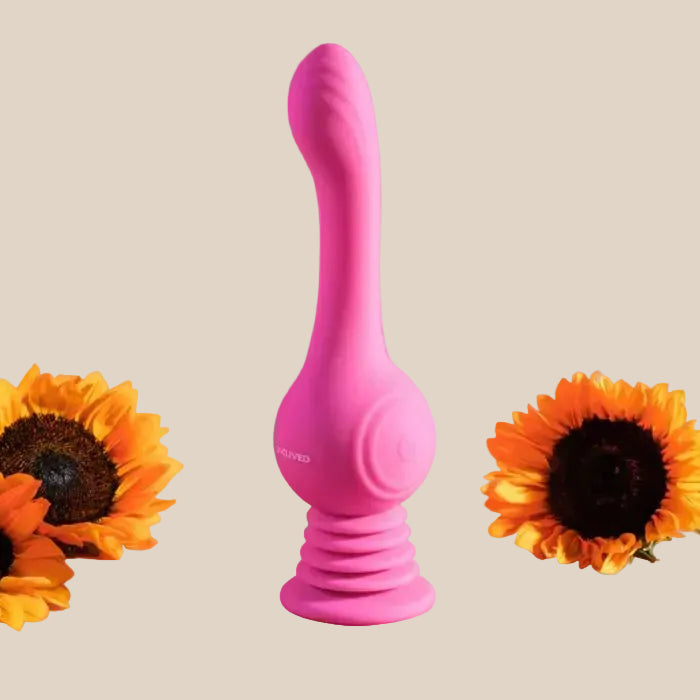 Pink silicone personal massager with curved design, displayed between two sunflowers on beige background