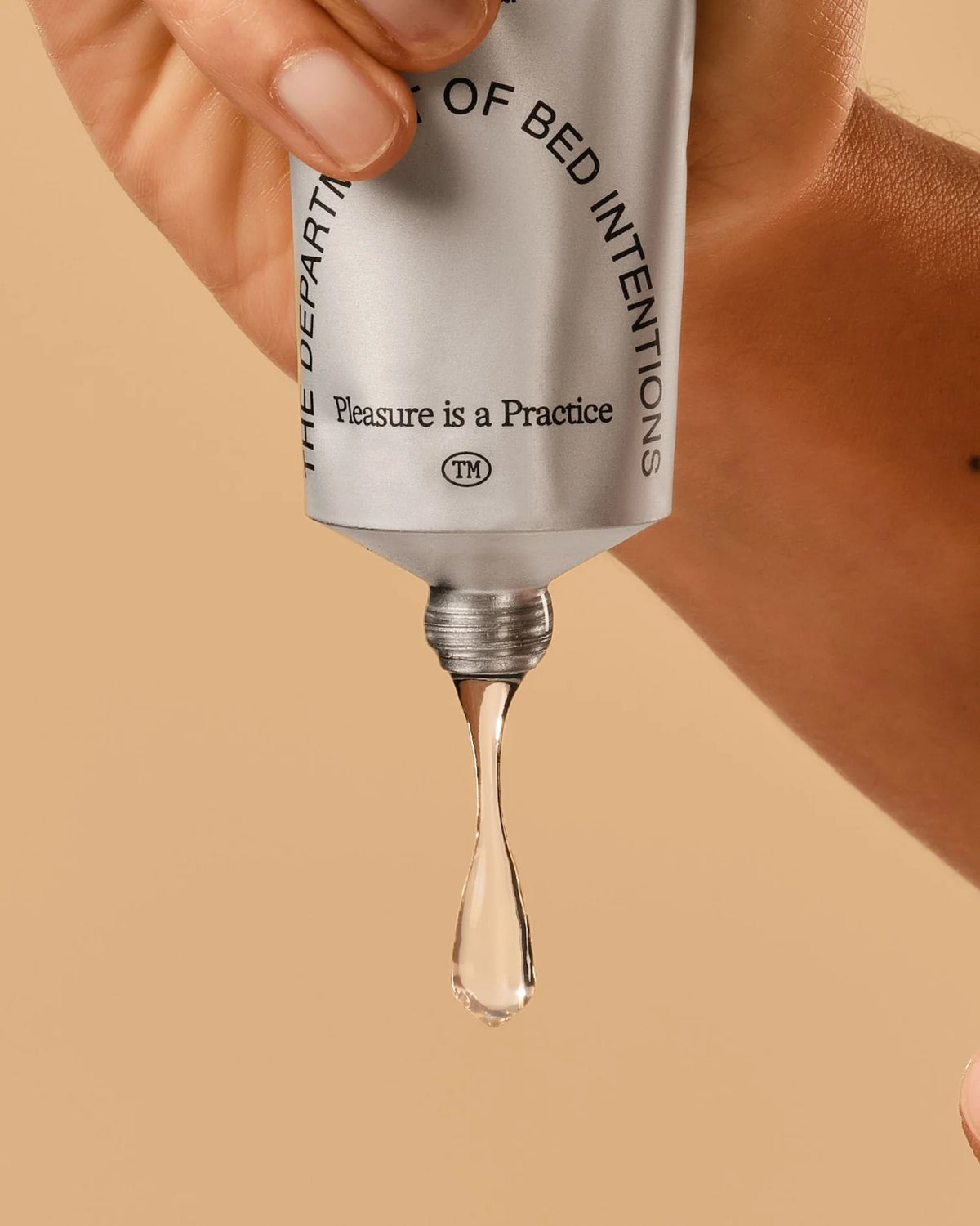 Hand squeezing clear lubricant gel from silver tube with 'Pleasure is a Practice' text