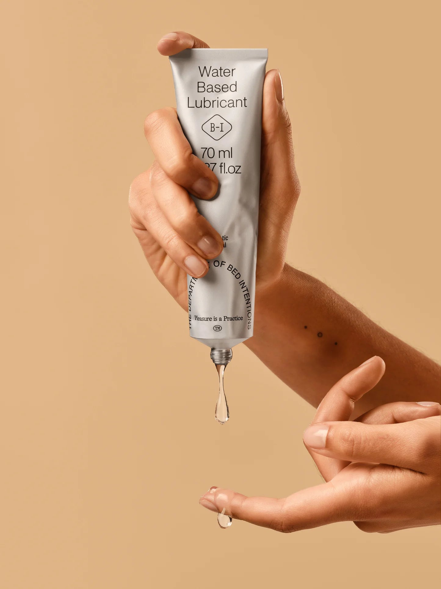 Hand squeezing clear water-based lubricant gel from a tube onto fingertips, beige background
