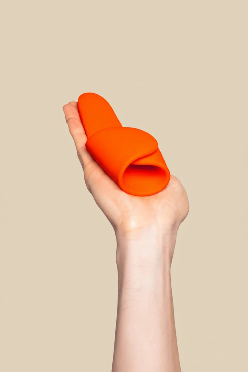 Hand holding an orange silicone inclusive handheld vibrator against a neutral background.
